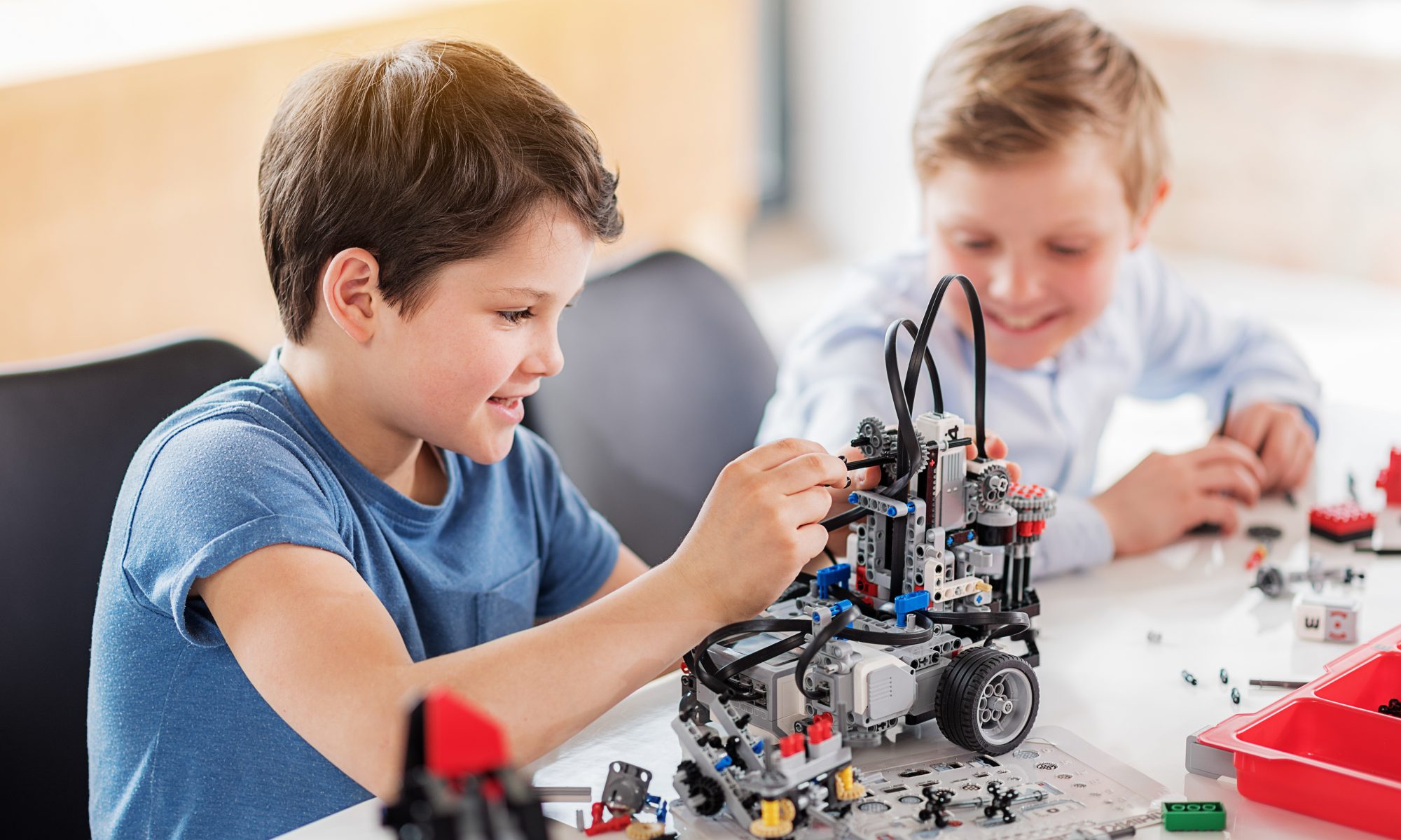 Interested male child is making robot, his friend looking at it with smile. They sitting near desk. Portrait